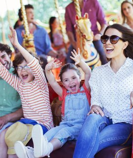 Menschen sitzen auf einem bunten Karussell im Vergnügungspark, dekorative Pferde und leuchtende Farben im Hintergrund sorgen für fröhliche Stimmung. | © Gettyimages.com/Caiaimage/Paul Bradbury
