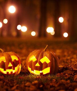 Zwei leuchtende Kürbisse mit geschnitzten Gesichtern auf einem mit Herbstlaub bedeckten Boden. Im Hintergrund sind verschwommene Lichter und Bäume zu sehen, was auf eine nächtliche Szene in einem Park oder Wald hindeutet. Die warmen Lichtakzente der Kürbisse schaffen eine stimmungsvolle, herbstliche Halloween-Atmosphäre. | © Gettyimages.com/sandsun