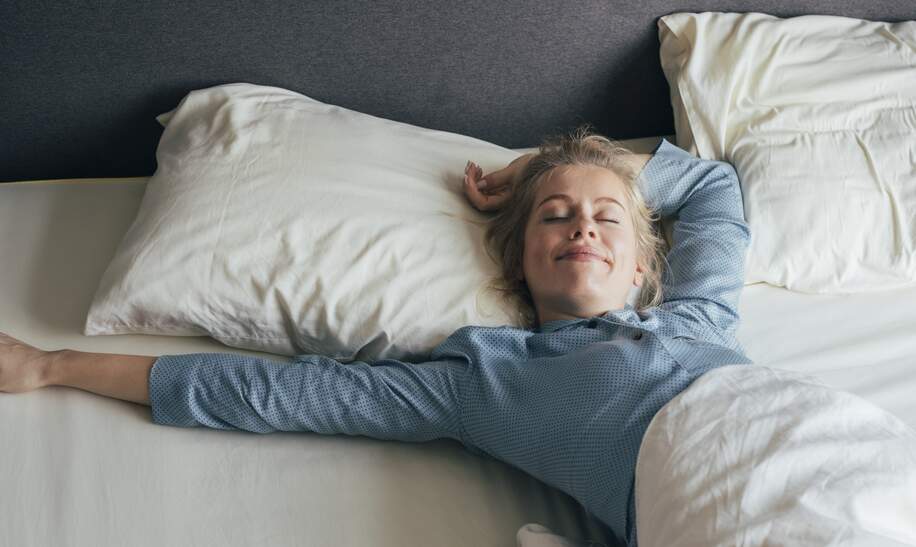 Laechelnde junge Frau, die an einem sonnigen Morgen ihre Arme im Bett streckt | © Gettyimages.com/FreshSplash