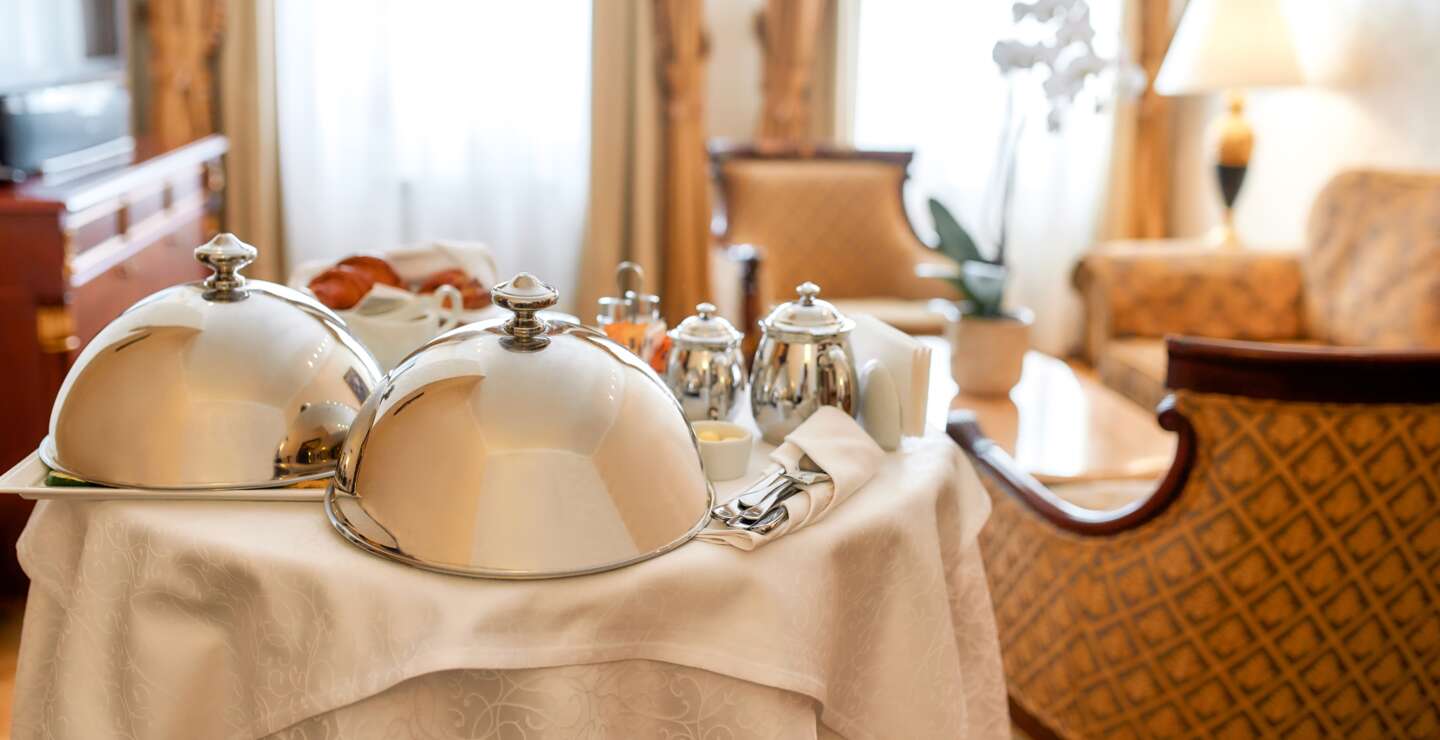 Luxurioeser Zimmerservice. Fruehstueck im luxurioesen Hotelzimmer vom Kellner geliefert. Mahlzeiten unter silberner Cloche. Gastfreundschaft und Urlaubskonzept. | © Gettyimages.com/LanaStock