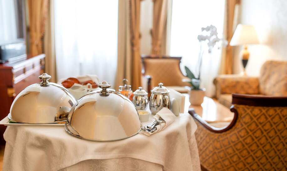 Luxurioeser Zimmerservice. Fruehstueck im luxurioesen Hotelzimmer vom Kellner geliefert. Mahlzeiten unter silberner Cloche. Gastfreundschaft und Urlaubskonzept. | © Gettyimages.com/LanaStock