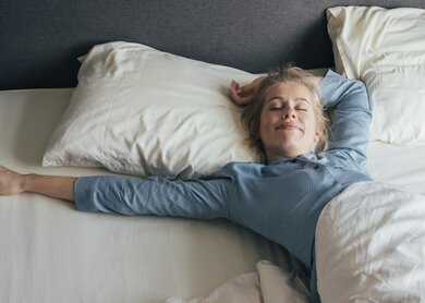 Laechelnde junge Frau, die an einem sonnigen Morgen ihre Arme im Bett streckt | © Gettyimages.com/FreshSplash