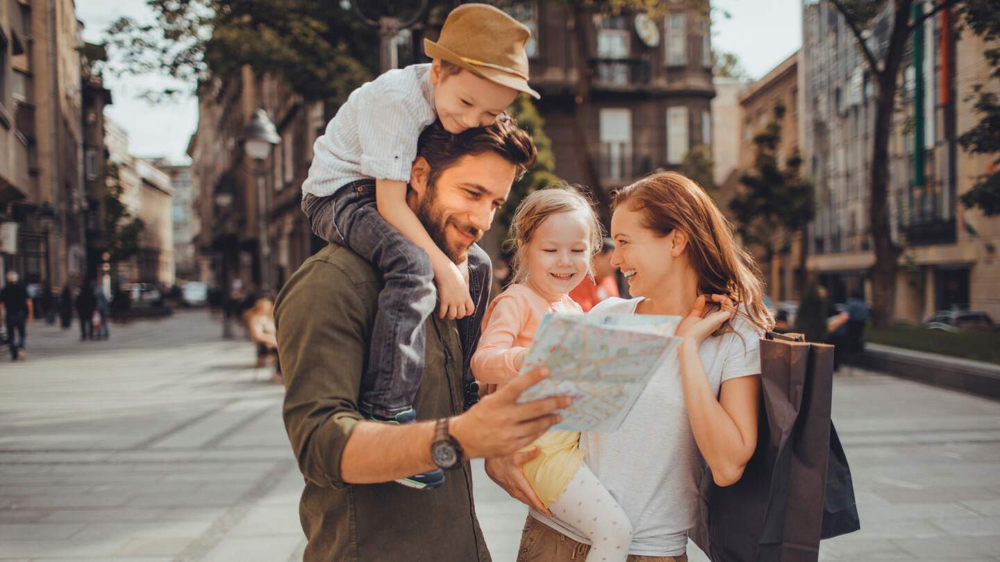 Familie, bestehen aus Mutter, Vater, Sohn und Tochter, gehen in der Stadt spazieren. Mutter trägt Tochter auf dem Arm, Sohn sitzt bei Vater auf den Schultern. | © Gettyimages.com/Geber86