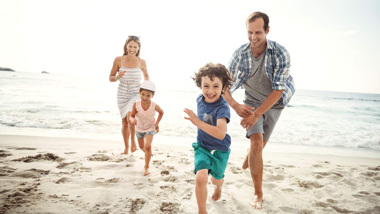 Familie läuft barfuß am Sandstrand entlang, im Hintergrund sind Meer, Wellen und heller Himmel zu sehen. | © GettyImages.com/shapecharge