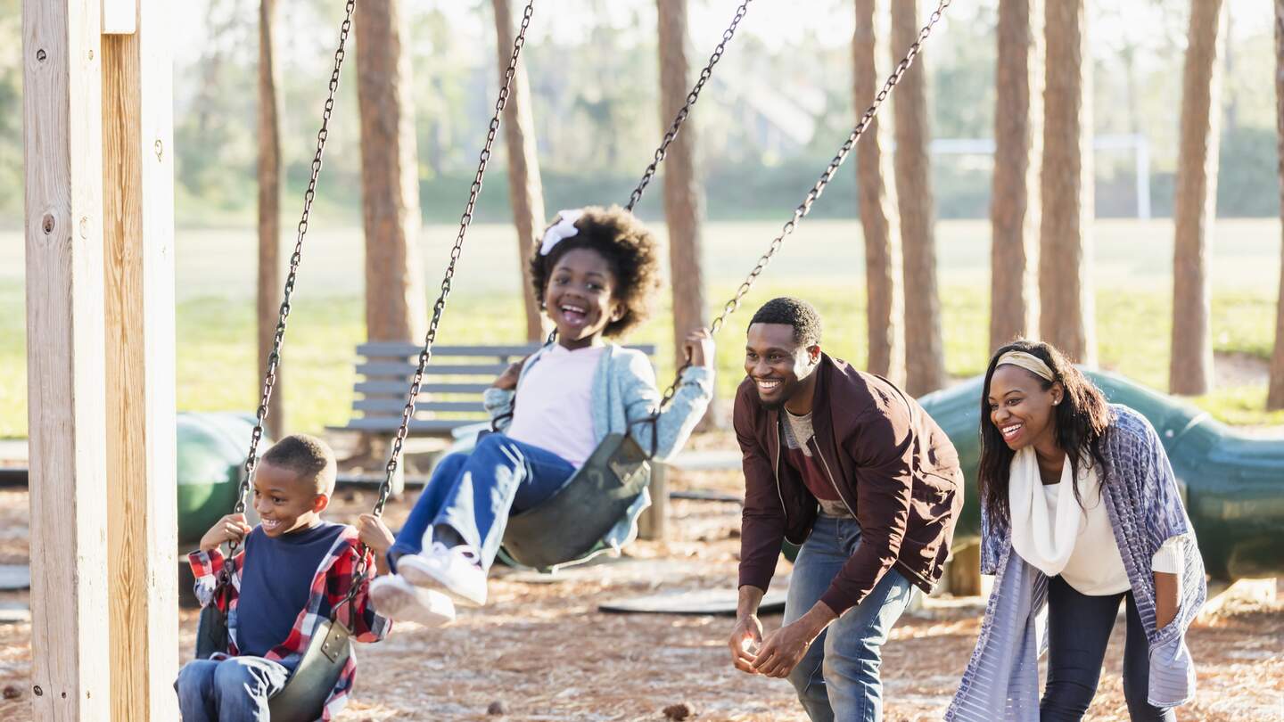 Eine Familie, die zusammen in einem Park spielt und Spass hat. Die Kinder, ein 7-jaehriger Junge und ein 5-jaehriges Maedchen, sind auf einer Schaukel und ihre Eltern schubsen ihre Schaukeln an. | © Gettyimages.com/Kali Nine LLC