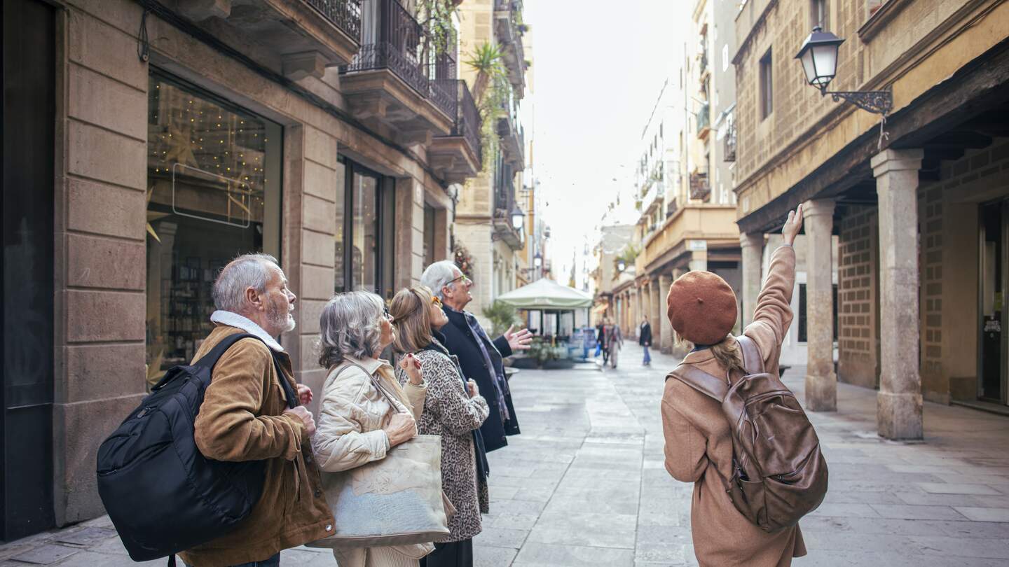 Eine Gruppe älterer Menschen steht in einer engen Altstadtstraße mit historischen Gebäuden und Balkonen, während eine Person auf architektonische Details zeigt. | © Gettyimages.com/DarioGaona