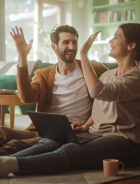 Paar benutzt Laptopund gibt sich ein High-Five, während sie im Wohnzimmer auf dem Fussboden sitzen | ©  Gettyimages.com/gorodenkoff
