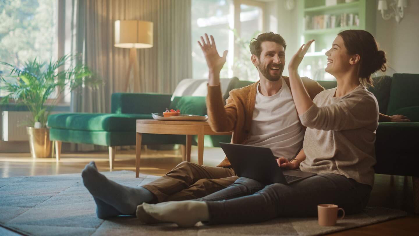 Paar benutzt Laptopund gibt sich ein High-Five, während sie im Wohnzimmer auf dem Fussboden sitzen | ©  Gettyimages.com/gorodenkoff