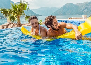 Paar hat Spass auf einer Luftmatratze im Pool mit einer bergigen Landschaft im Hintergrund | © Gettyimages.com/DaniloAndjus