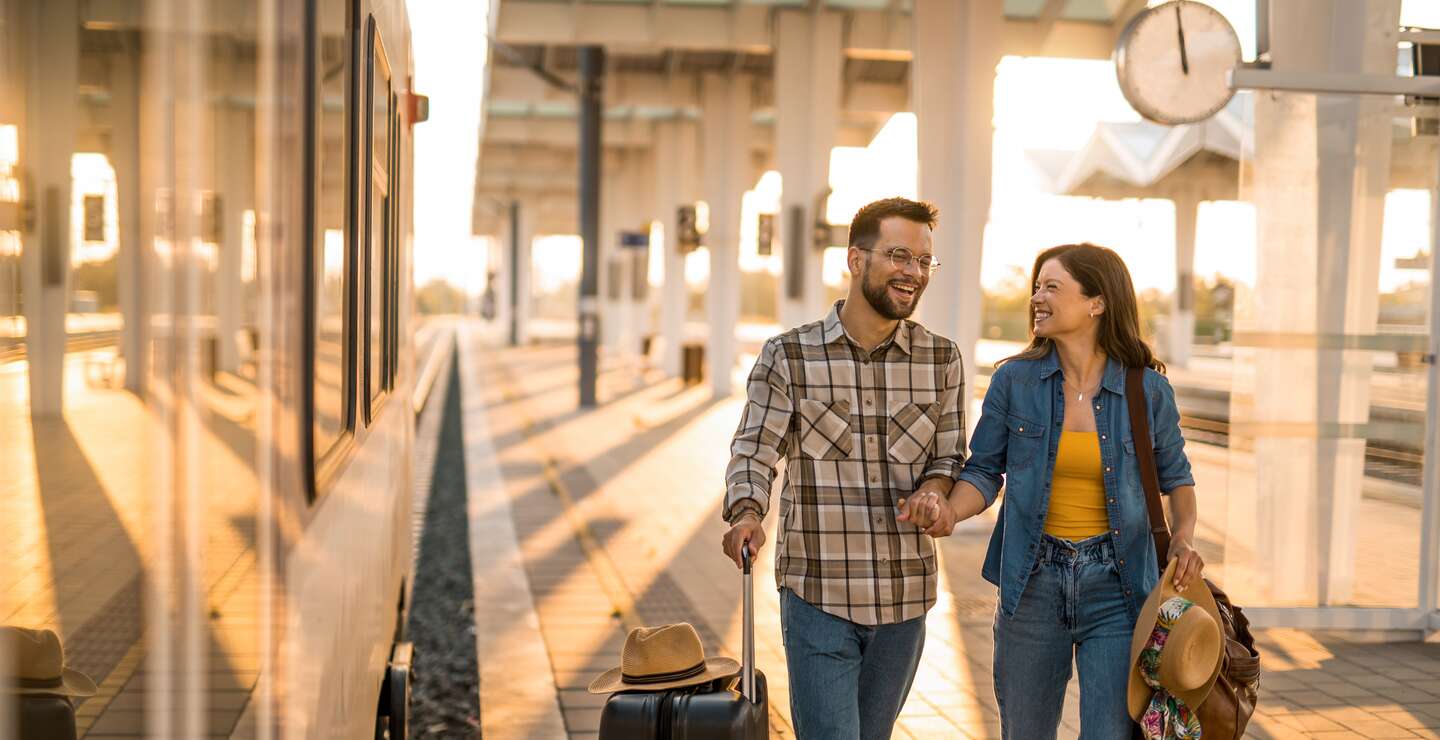 Mann und Frau laufen lachend einen Bahnsteig mit Koffer und Reisetasche entlang | © Gettyimages.com/DjelicS
