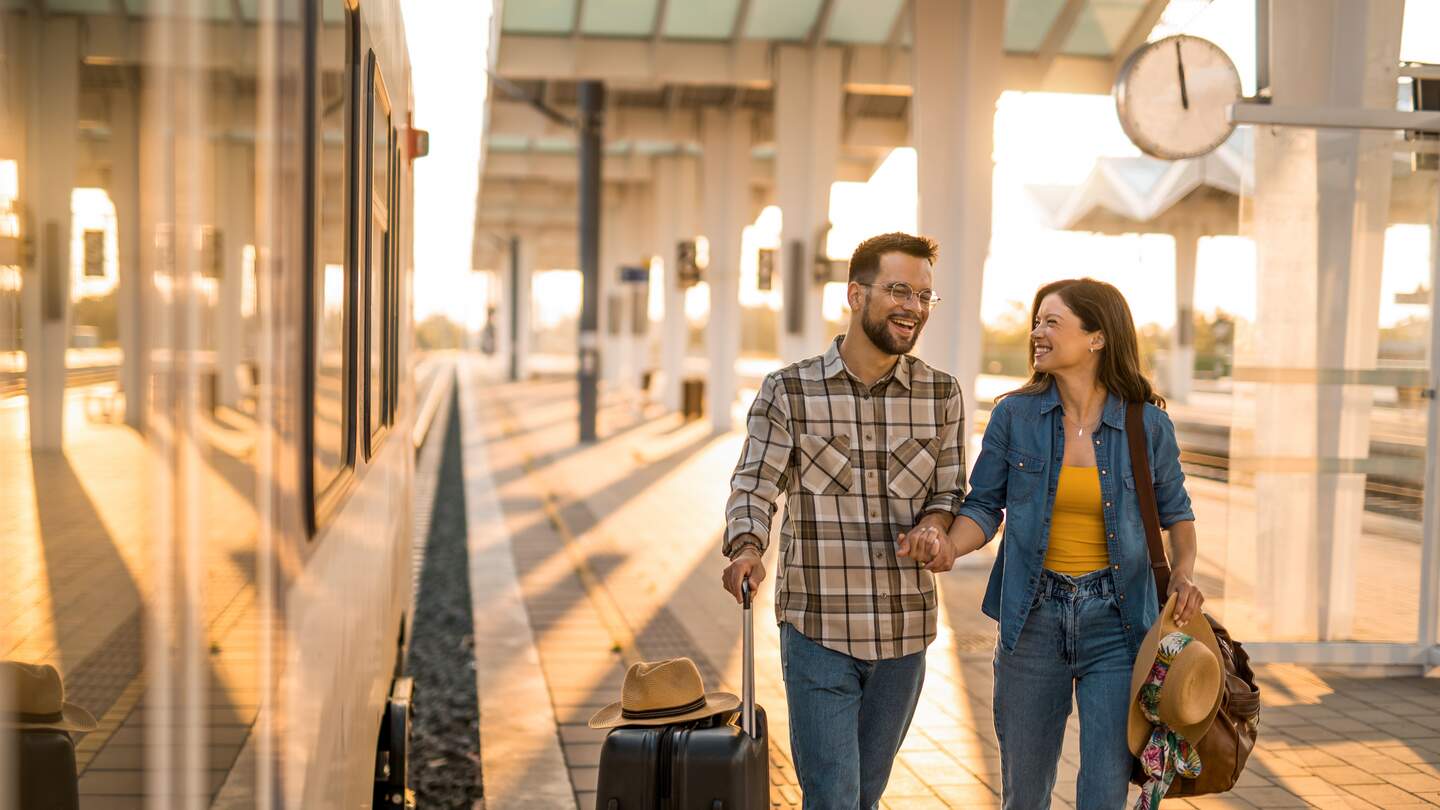 Mann und Frau laufen lachend einen Bahnsteig mit Koffer und Reisetasche entlang | © Gettyimages.com/DjelicS