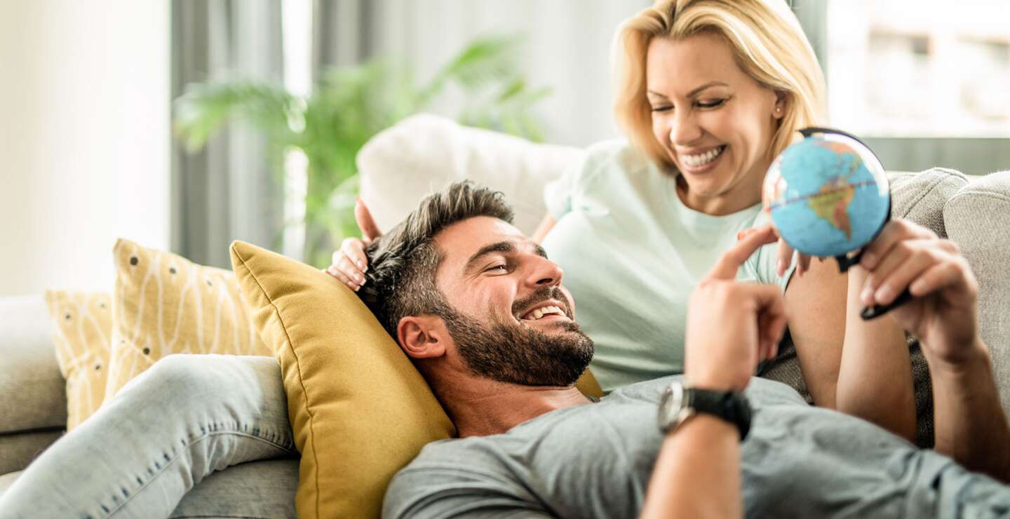 Glueckliches junges Paar plant ihre neue Reise. Junger Mann entspannt sich mit seiner Freundin auf dem Sofa und blickt auf den Globus. | © GettyImages.com/DjelicS