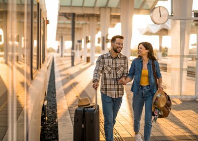 Mann und Frau laufen lachend einen Bahnsteig mit Koffer und Reisetasche entlang | © Gettyimages.com/DjelicS