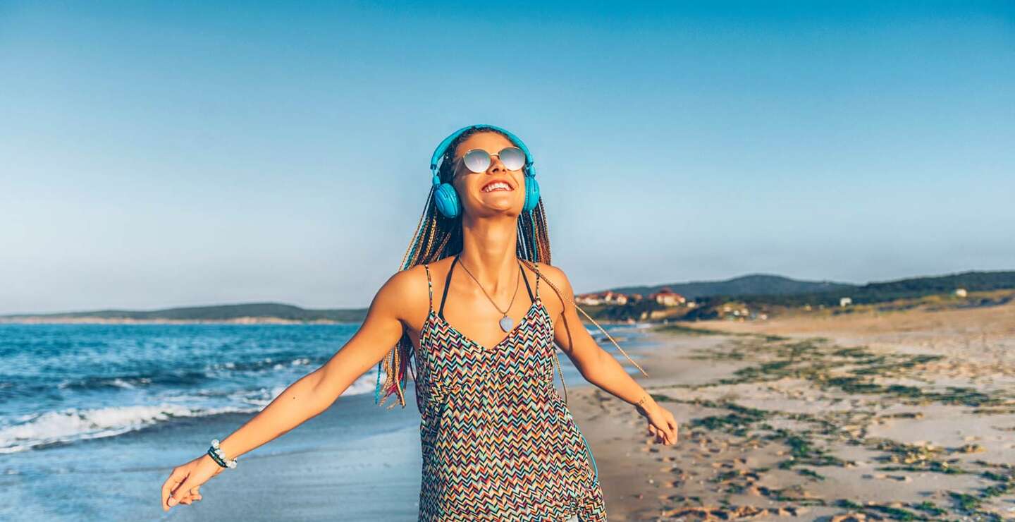 Froehliche junge Frau mit blauen Kopfhoerern, die am Strand Musik hört | © Gettyimages.com/Vladimir Vladimirov