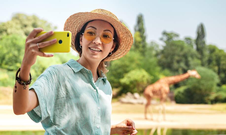 Studentin macht ein Selfie auf dem Smartphone, während eine Giraffe aus dem See trinkt | © Gettyimages.com/frantic00