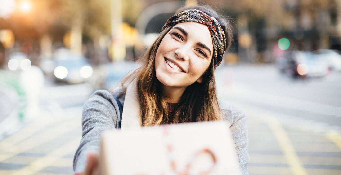 Junge Frau, die in der Stadt an einer Strasse ein Geschenk ueberreicht. | © GettyImages.com/	Tempura