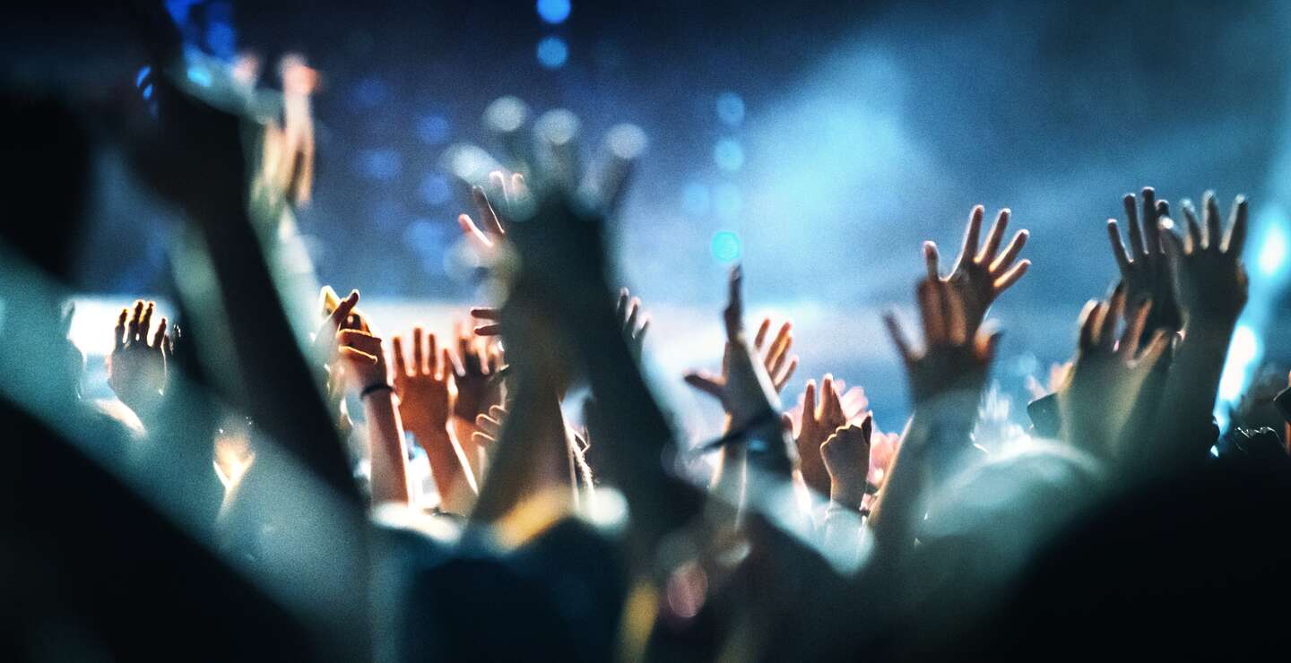 Rueckansicht einer grossen Gruppe von Menschen, die eine Konzertauffuehrung geniessen. Es gibt viele Haende, die applaudieren und die Show aufnehmen. Mehrfarbige Laser und Spotlichter feuern von der Buehne.  | © Gettyimages.com/gilaxia
