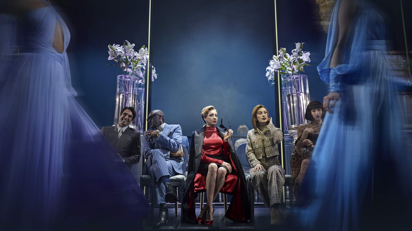 Frau sitzt im roten Kleid auf einem Stuhl und sieht mit anderen Menschen eine Modenschau an | © Stage Entertainement, Matt Crockett