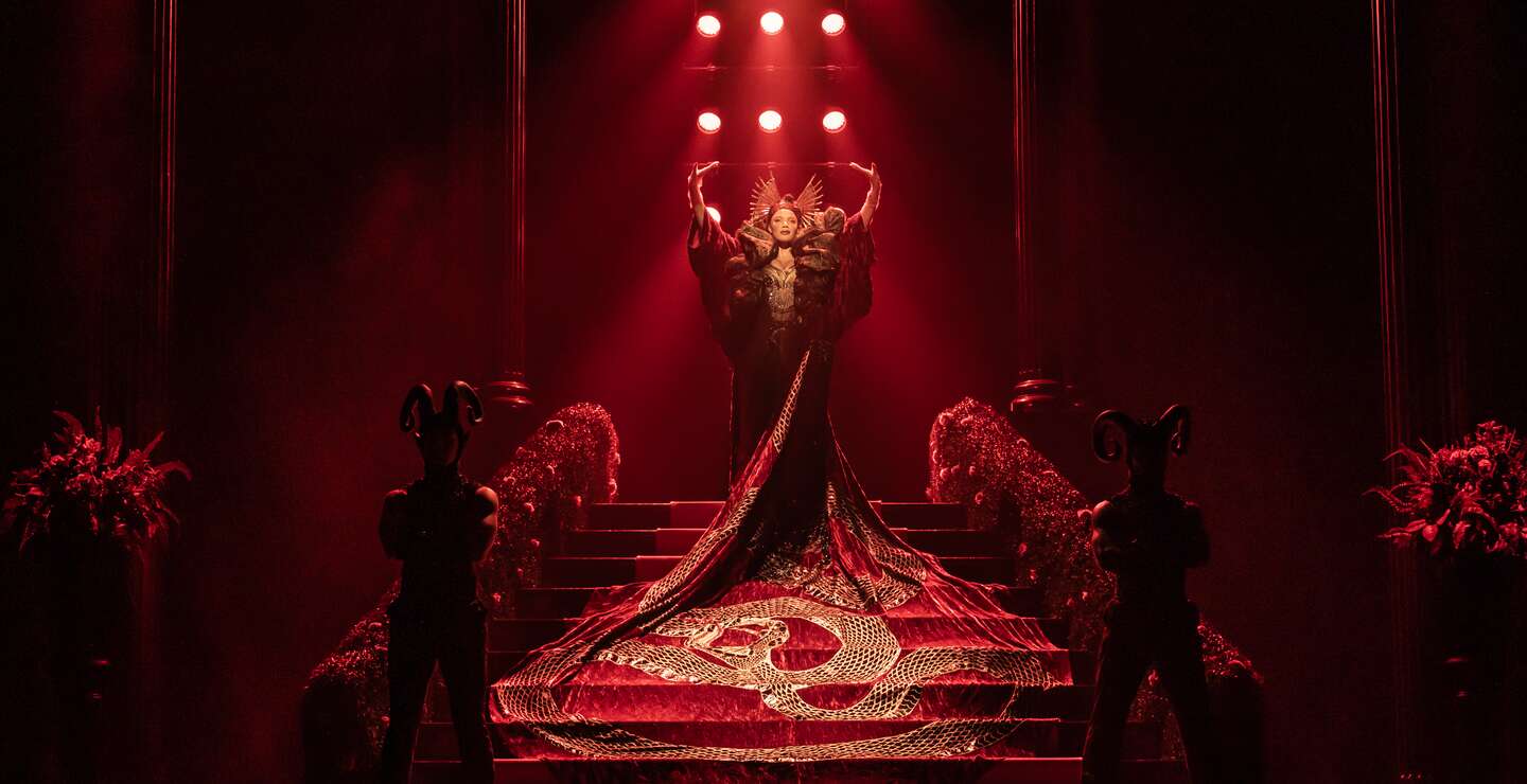 Frau steht in einem rotem Kleid mit  langer Schleppe auf einer roten Treppe | © Stage Entertainement, Matt Crockett