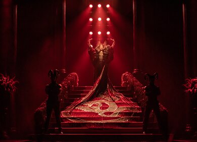Frau steht in einem rotem Kleid mit  langer Schleppe auf einer roten Treppe | © Stage Entertainement, Matt Crockett