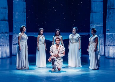 Disneys Hercules singend auf dem Knie im Vordergrund, Saenger in weissen Kleidern im Kreis herumstehend | © Stage Entertainment/Johan Persson 