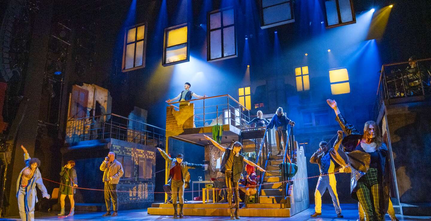 Wir sind am Leben-Ensemble performt auf mehrstöckigem Bühnenbild mit Treppen und Fenstern, dramatische Lichtstimmung in Blau und Gelb | © Stage Entertainement, Joern Hartmann