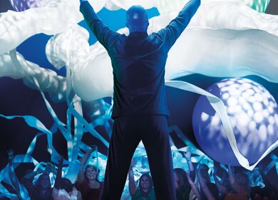 Einer der Kuenstler guckt zum Publikum und hebt die Haende, ueber dem Publikkum schweben Ballons | © Stage Entertainment