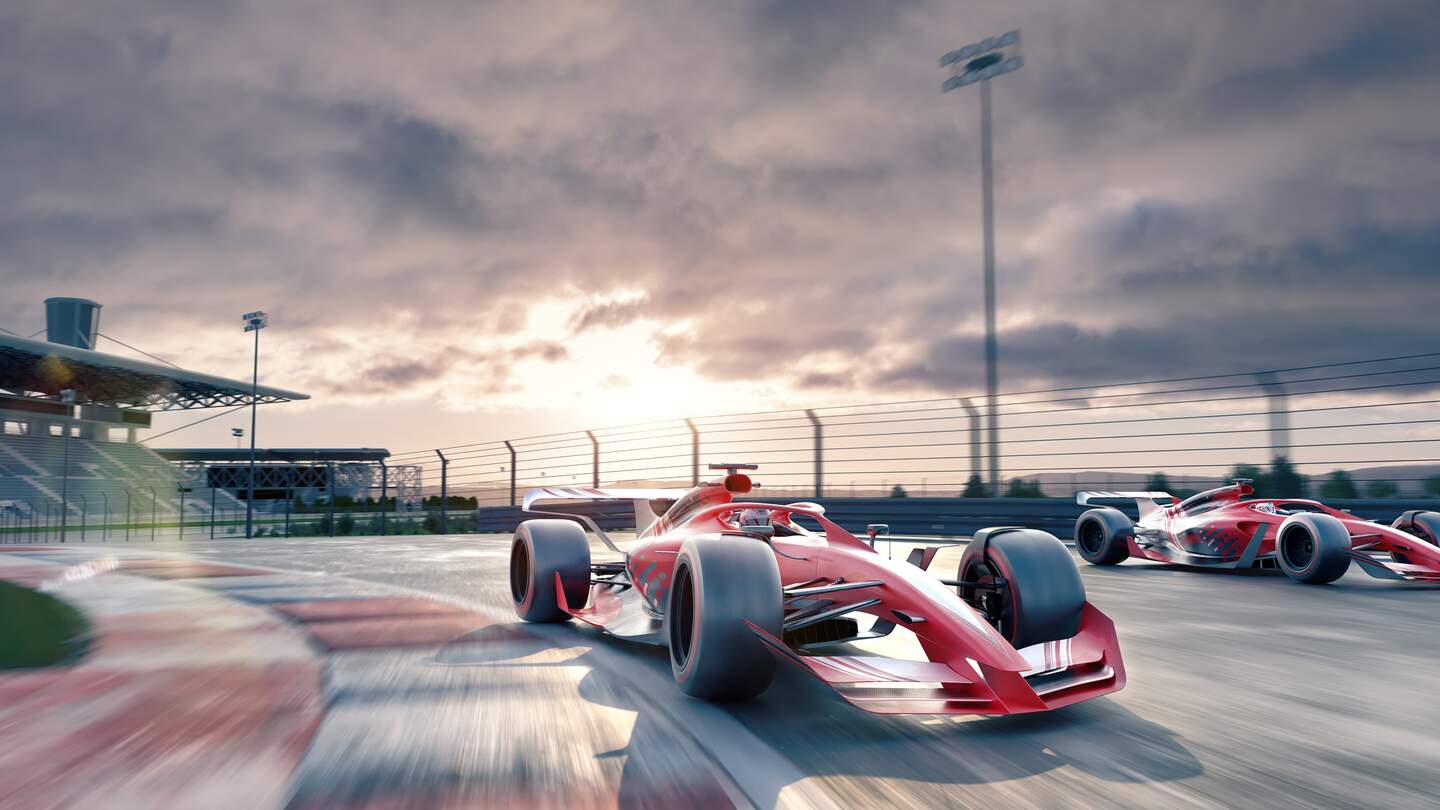 Zwei generische Rennwagen, die sich mit hoher Geschwindigkeit bewegen, bei Sonnenaufgang/Sonnenuntergang unter bewoelktem Himmel um eine Ecke einer Rennstrecke fahren, mit leeren Zuschauerraengen im Hintergrund. Mit Bewegungsunschaerfe zu Hintergrund und Lens Flare. | © Gettyimages.com/peepo