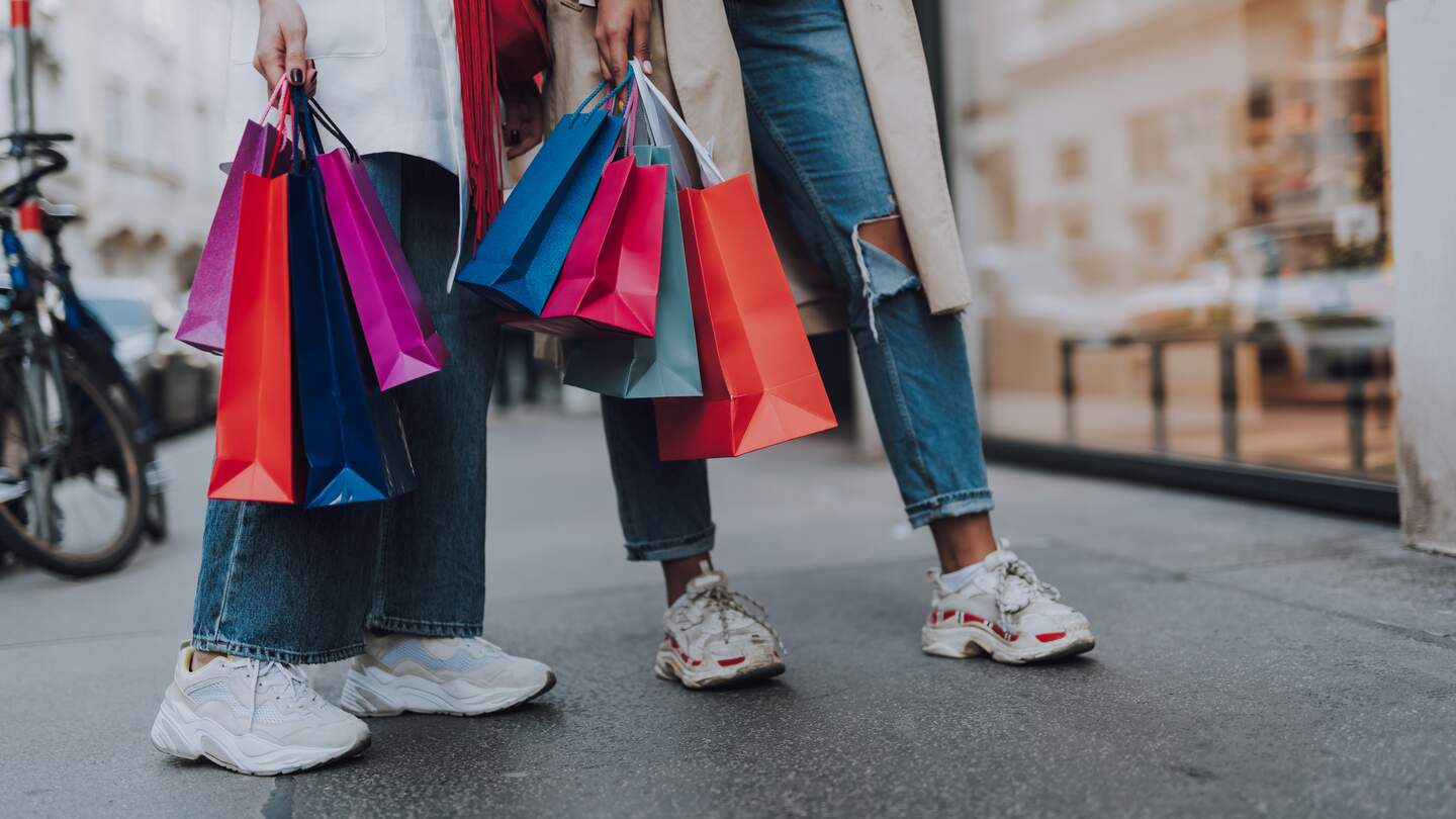 Nahaufnahme von Beinen zweier Damen in Jeans und Turnschuhen mit bunten Einkaufstaschen | © Gettyimages.com/Yaroslav Astakhov