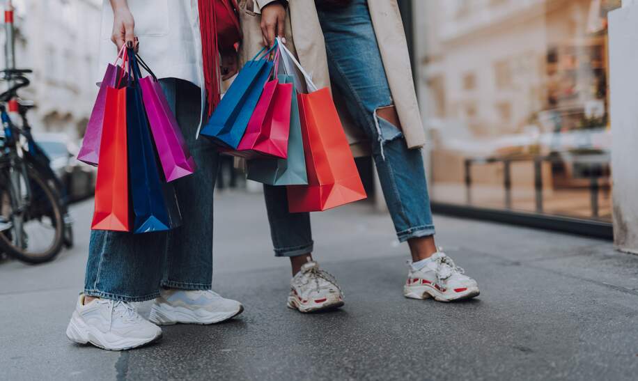 Nahaufnahme von Beinen zweier Damen in Jeans und Turnschuhen mit bunten Einkaufstaschen | © Gettyimages.com/Yaroslav Astakhov