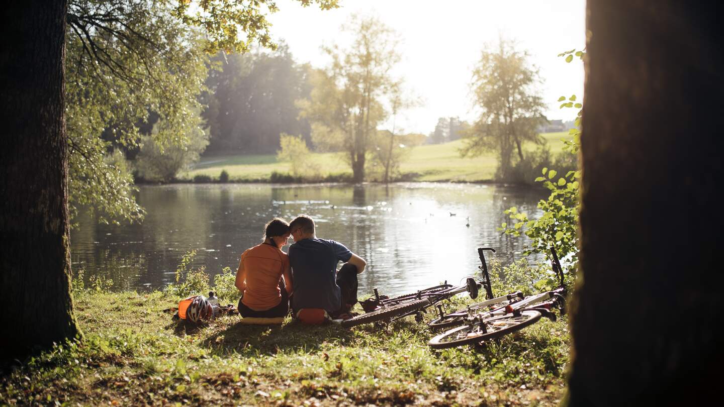 Junges Paar sitzt am See und lehnt die Koepfe aneinander, daneben liegen ihre Fahrraeder auf dem Boden und die Sonne scheint auf dem See  mit dem Wald im Hintergrund | © GettyImages.com/AzmanL