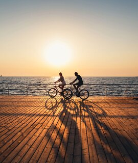 Ein junges Paar fährt zusammen Fahrrad auf einem Holddeck am Strand bei Sonnenaufgang  | © GettyImages.com/IB_photo