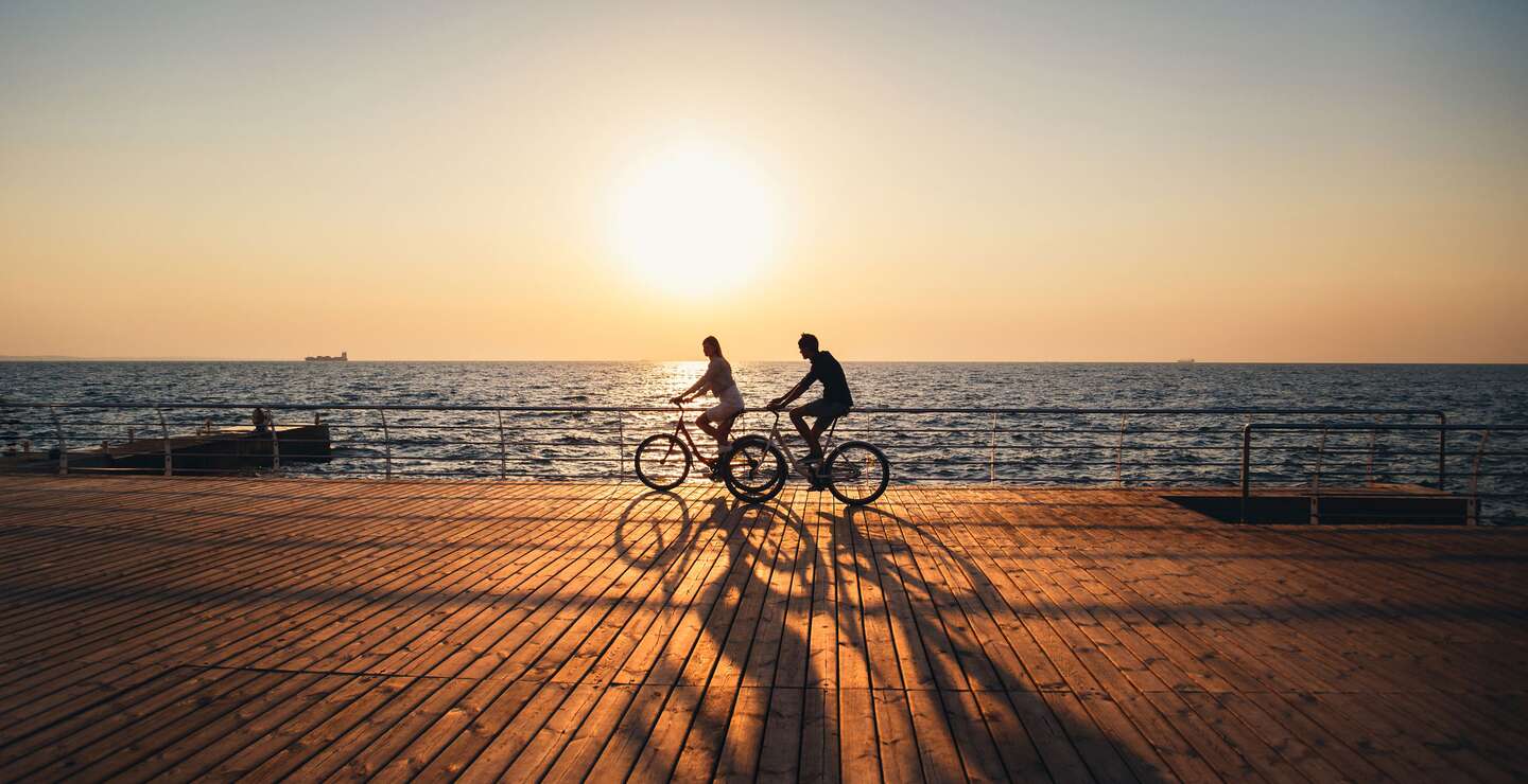 Ein junges Paar fährt zusammen Fahrrad auf einem Holddeck am Strand bei Sonnenaufgang  | © GettyImages.com/IB_photo