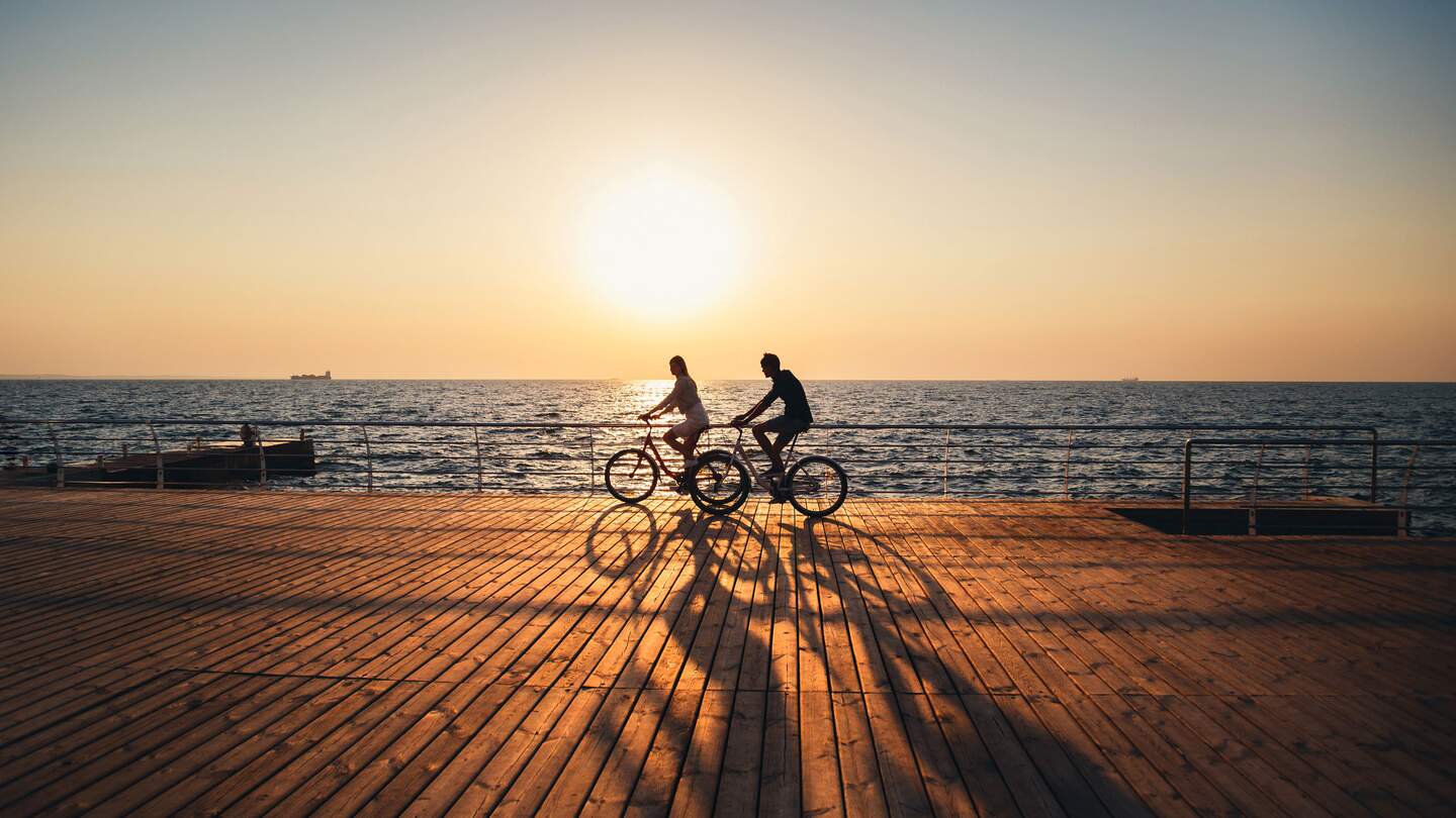 Ein junges Paar fährt zusammen Fahrrad auf einem Holddeck am Strand bei Sonnenaufgang  | © GettyImages.com/IB_photo