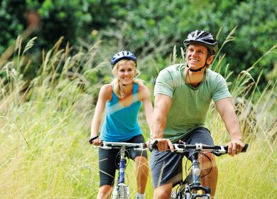 Paar fährt mit kurzer Kleidung im Feld mit Hohen Gräsern und Wald im Hintergrund Fahrrad | © GettyImages.com/warrengoldswain