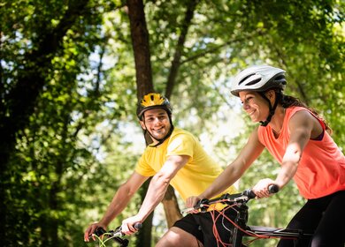 Das junge Paar faehrt im Sommer mit kurzer Kleidung auf einem Weg mit Baeumen | © GettyImages.com/microgen