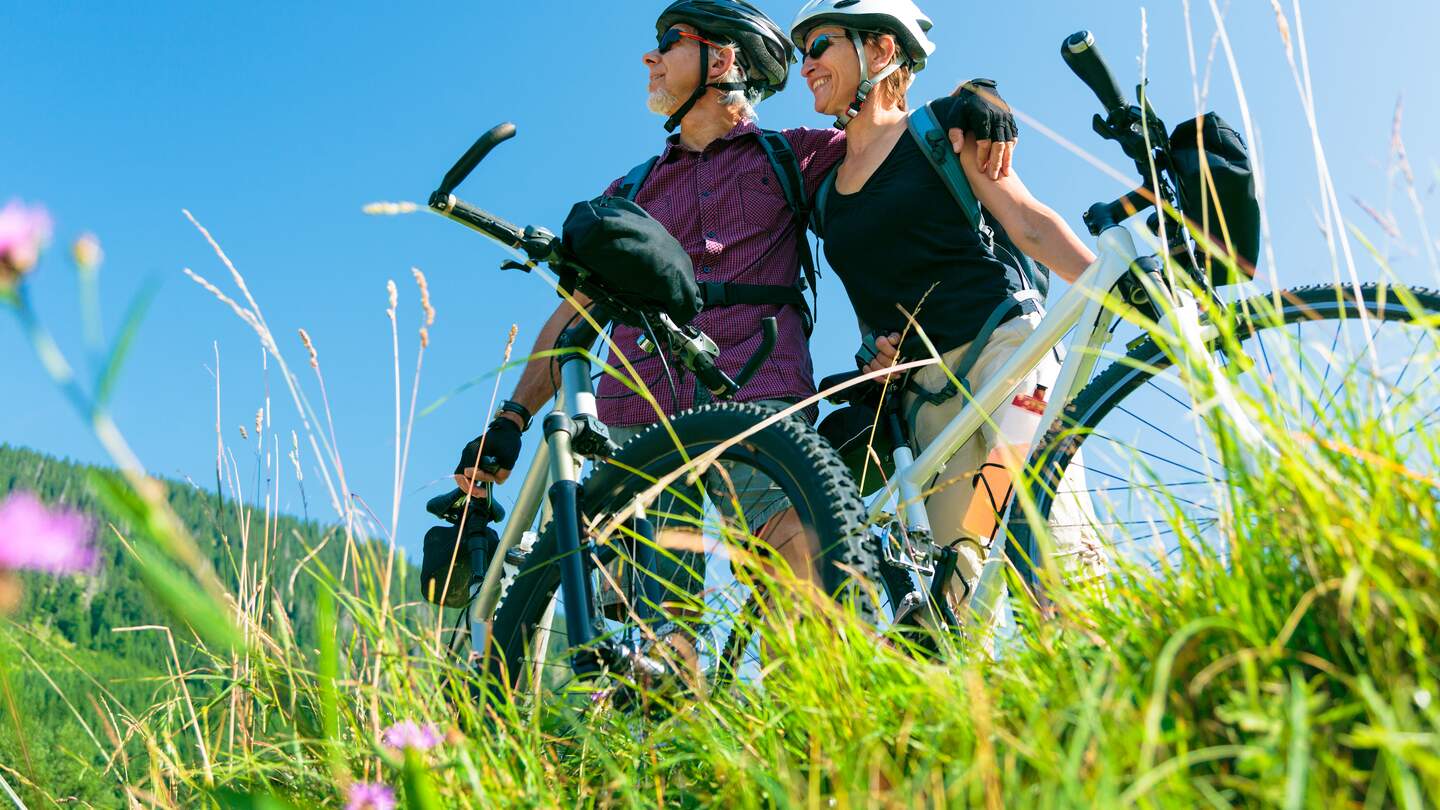 Ein Seniorenpaar steht mit Fahrraedern auf einer gruenen Wiese, schaut in die Ferne und ist aus der Untersicht zu sehen, der Himmel ist strahlend blau | © GettyImages.com/nullplus