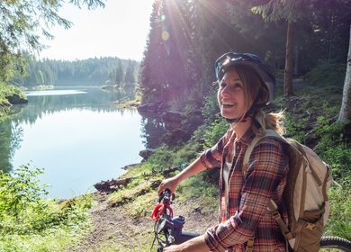 Frau auf dem Mountainbike mit Blick auf schönen Alpensee mit Sonnenschein | © GettyImages.com/Mystockimages