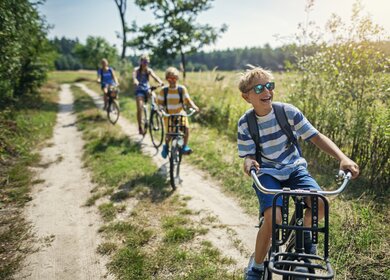 Drei Kinder fahren voraus auf dem Feldweg angrenzed an einen Wald und die Mutter ist verschwommen im Hintergrund zu sehen | © GettyImages.com/Imgorthand