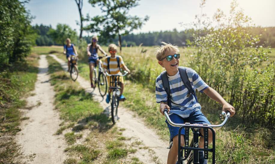 Drei Kinder fahren voraus auf dem Feldweg angrenzed an einen Wald und die Mutter ist verschwommen im Hintergrund zu sehen | © GettyImages.com/Imgorthand