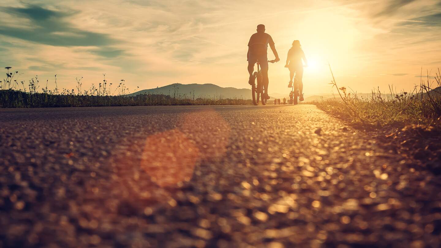 Paar auf Fahrraedern ist von Hinten zu sehen, waehrend sie Richtung Sonne auf einem Weg im Feld mit Bergen fahren und die sonne reflektiert | © GettyImages.com/Solovyova