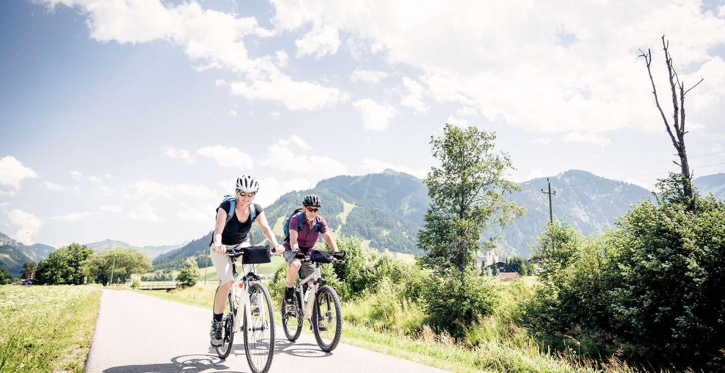 Senioren-Paar faehrt mit Fahrraedern ueber einen Feldweg mit einer Berglandschaft hinter ihnen | © Gettyimage/nullplus