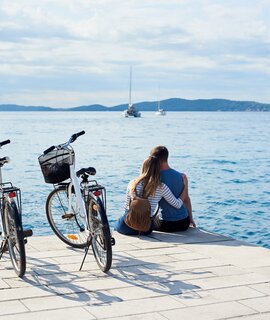 Man und Frau mit Rucksack sitzen umarmt auf Fahrraedern, beobachten schwimmende Kreuzfahrtschiffe in klaren blauen Meer.  | © Gettyimages.com/anatoliy_gleb