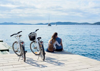 Man und Frau mit Rucksack sitzen umarmt auf Fahrraedern, beobachten schwimmende Kreuzfahrtschiffe in klaren blauen Meer.  | © gettyimages.com/anatoliy_gleb
