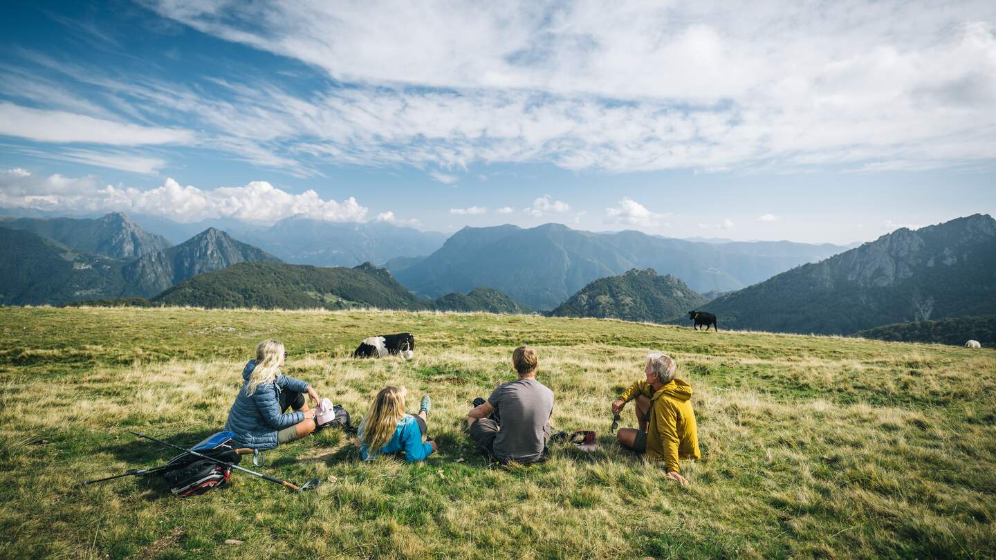 Mehrere Wandernde sitzen auf einer Almwiese und blicken auf ein weitläufiges Bergpanorama unter blauem Himmel mit Wolken; im Hintergrund grasen Kühe. | © GettyImages.com/	AscentXmedia