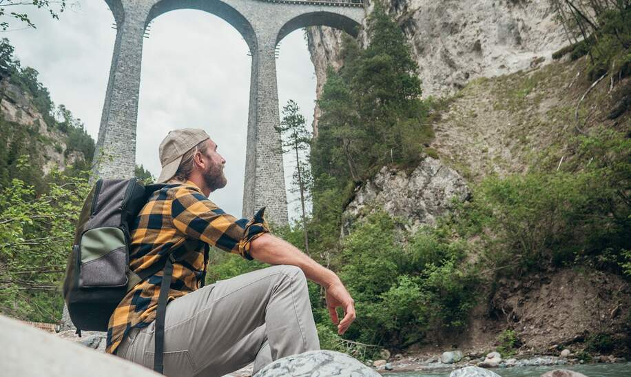Ein Mann sitzt auf Felsen am Fluss und schaut auf eine grosse Steinbogenbruecke in der Ferne | © Gettyimages.com/Mystockimages