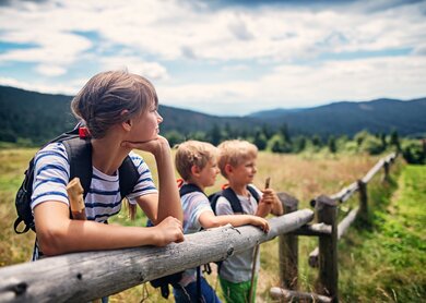 Drei Kinder im Alter von 10 und 7 Jahren vergnügen sich beim Wandern auf den Feldern und Wiesen. Sanft geneigte Hügel, die von Mischwald bedeckt sind, sind im Hintergrund zu sehen. Kinder lehnen sich an einen Holzzaun.  | © Gettyimages.com/Imgorthand