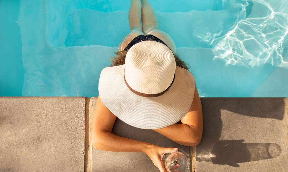 Hochwinkelansicht einer Frau im Bikini und Sonnenhut, die in einem Schwimmbad sitzt und ein Glas Wein trinkt | © Gettyimages.com/NickyLloyd