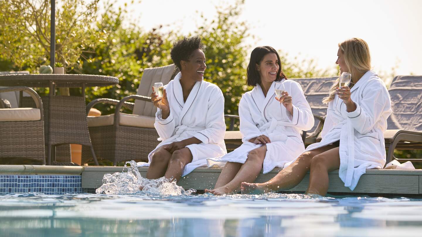 Gruppe von Freundinnen, die Bademaentel tragen, sitzen mit den Fuessen im Pool und trinken Champagner am Spa-Tag | © Gettyimages.com/monkeybusinessimages