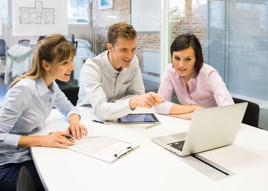 Drei Personen sitzen an einem Tisch in einem gut beleuchteten Raum und besprechen Dokumente. Im Hintergrund sind architektonische Zeichnungen zu sehen. | © Gettyimages.com/LDProd 